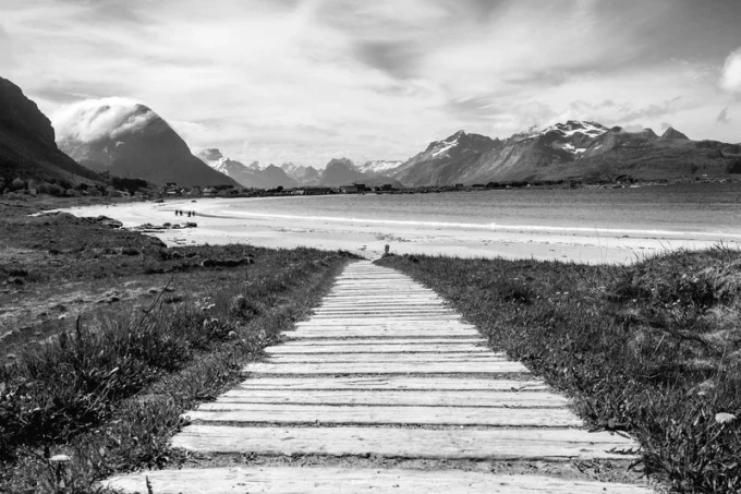 Beautiful wall mural of a scenic road leading to a lakeside village, featuring a wooden path through a meadow.