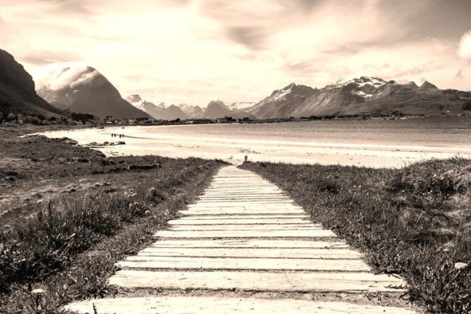 Beautiful wall mural of a scenic road leading to a lakeside village, featuring a wooden path towards the mountains.