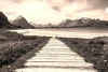 Beautiful wall mural of a scenic road leading to a lakeside village, featuring a wooden path towards the mountains.